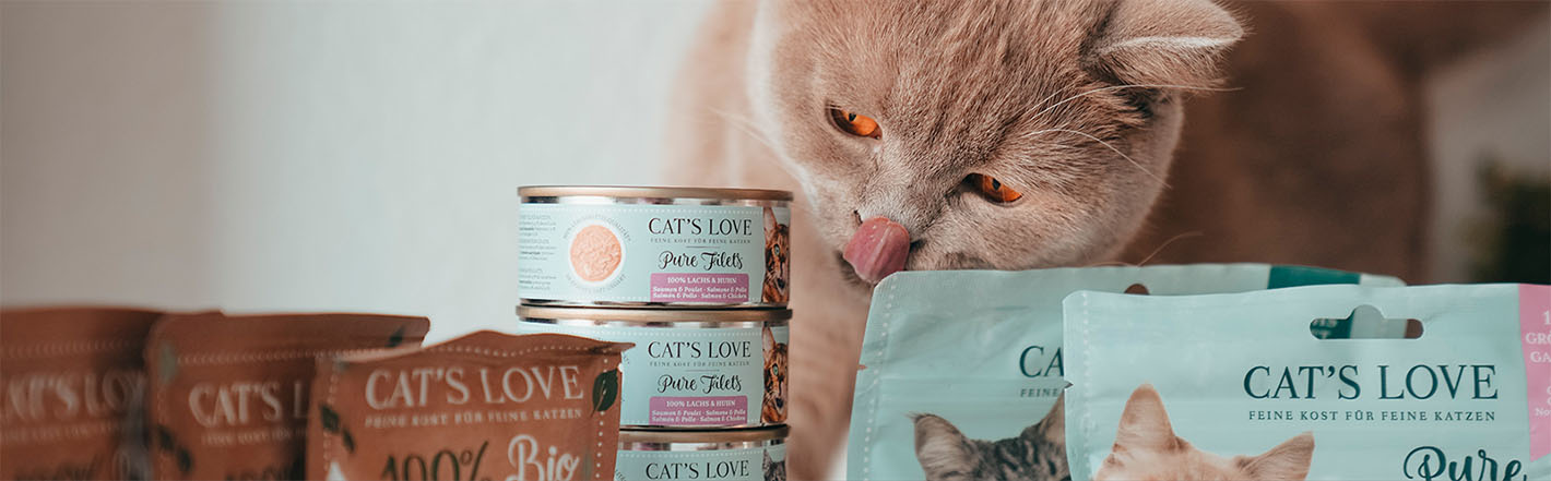 Un gato sentado detrás de diferentes productos CAT'S LOVE y lamiéndose la boca.